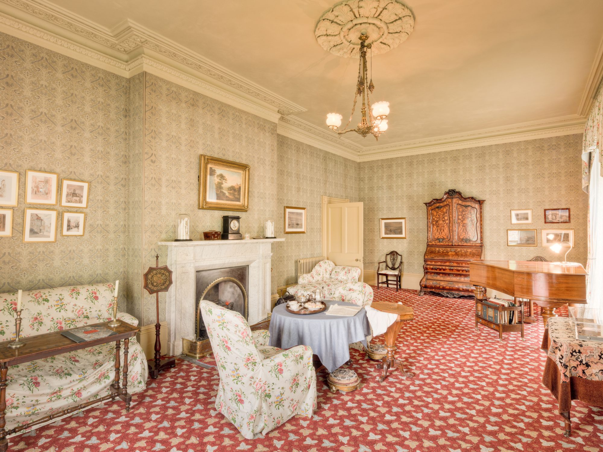 Traditional drawing room papered in a small-scale pattern beneath deep crown molding, with a marble fireplace, chintz armchairs and a brass chandelier