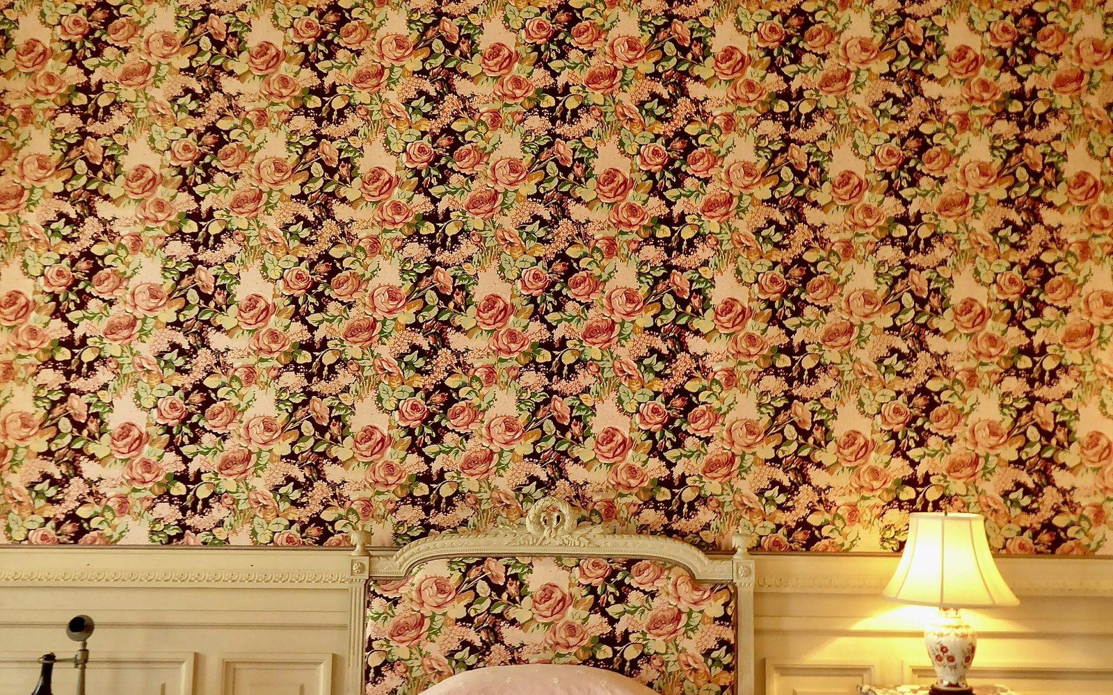 Bedroom papered in a dense rose-and-lilac floral above cream paneled wainscoting, with a matching upholstered headboard and a lit table lamp