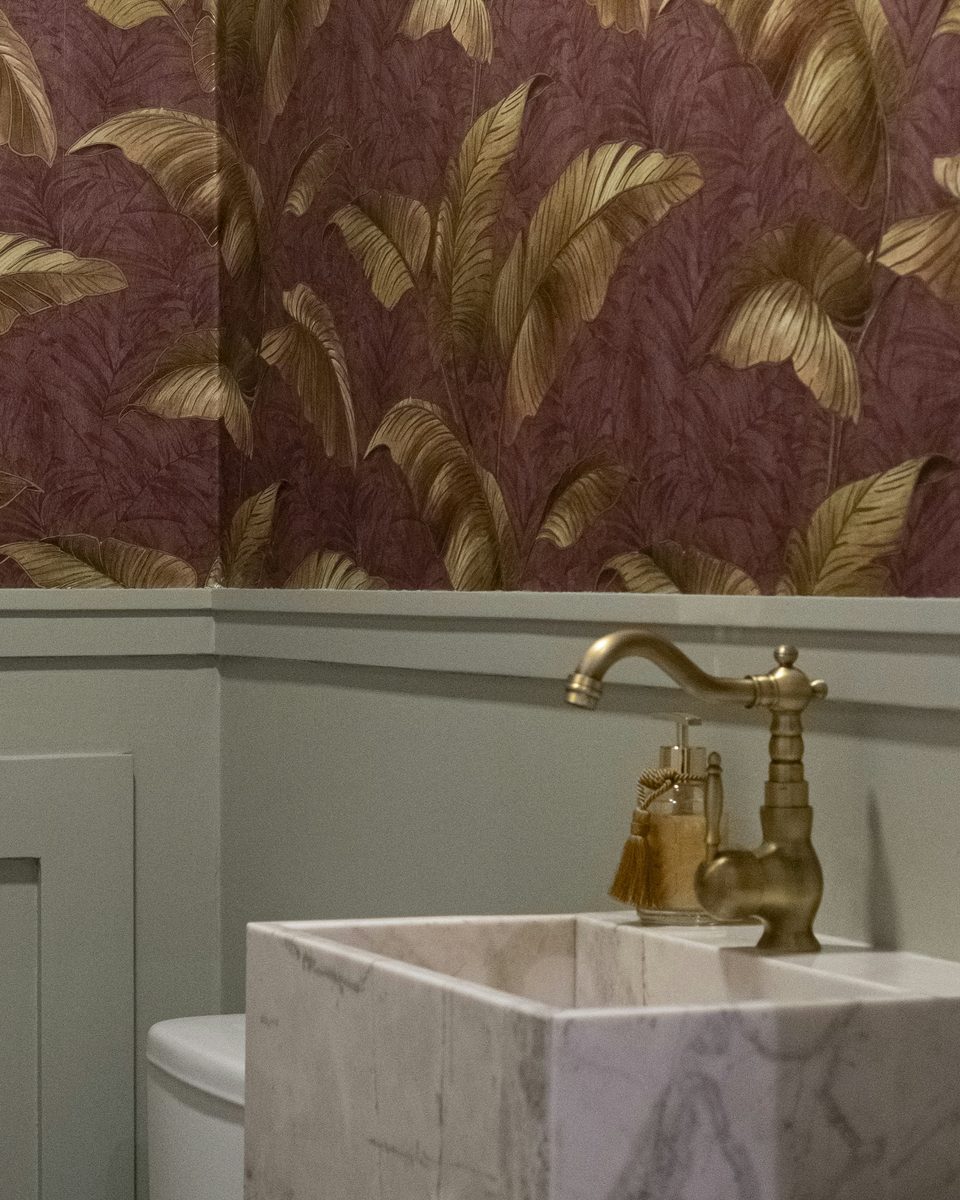 Powder room with gold palm-leaf wallpaper on a plum ground above sage paneling, a brass faucet and a marble pedestal sink