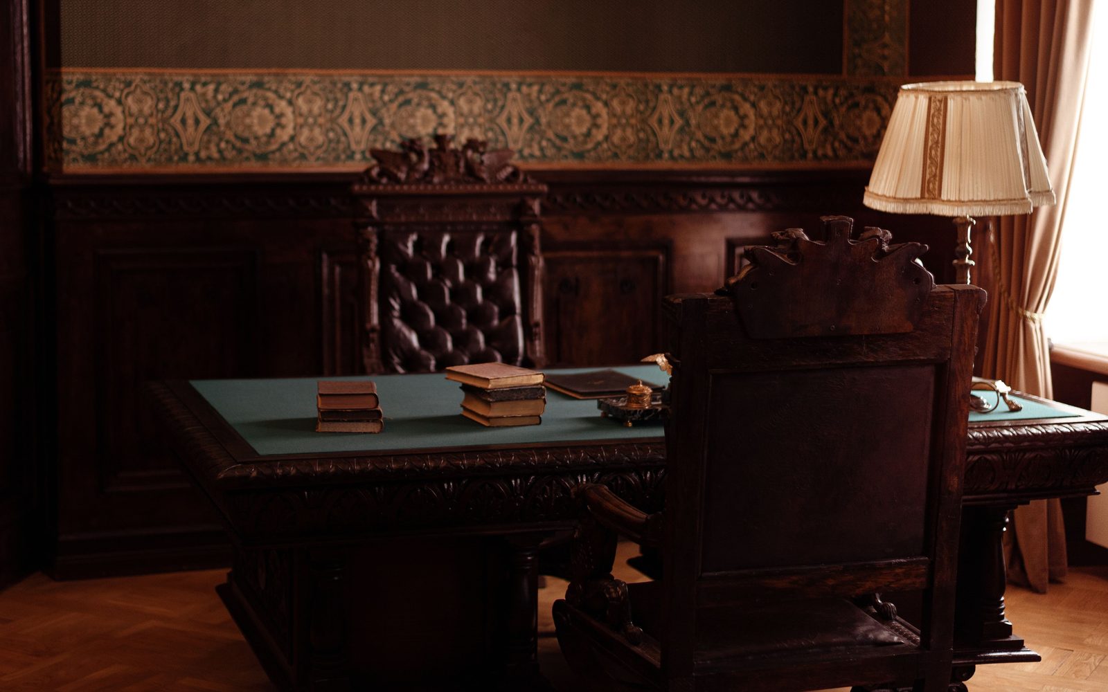 Study with dark wood paneling topped by an ornamental wallpaper border, a carved desk with a green leather top and a pleated lamp