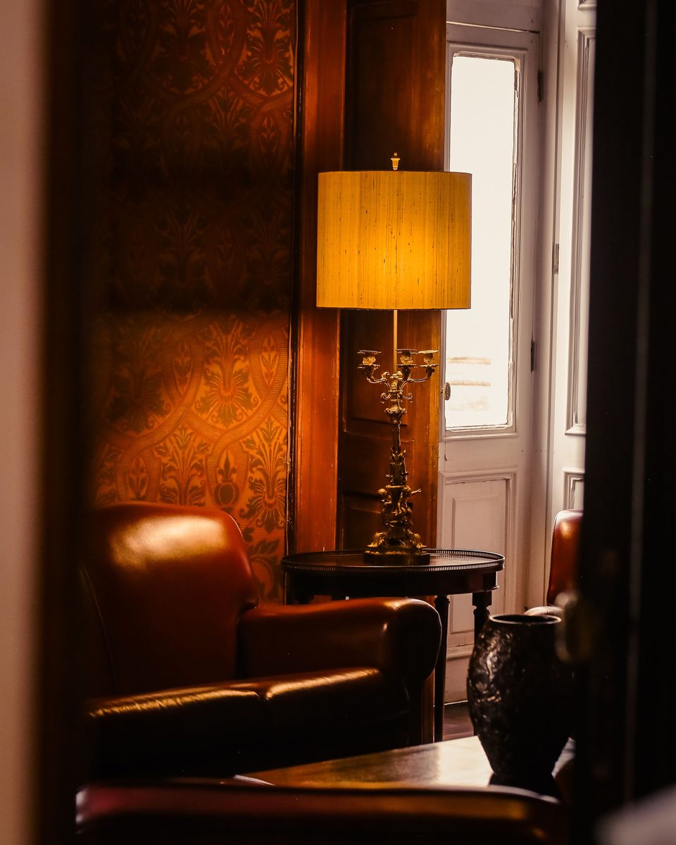Warm lounge corner with a rust-toned patterned wallpaper, a leather armchair and a brass table lamp beside a tall window