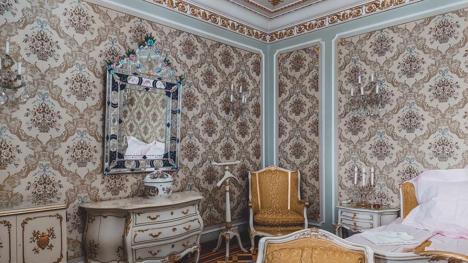 Bedroom with damask-patterned wallpaper set inside painted wall panels, a gilded mirror over a carved dresser